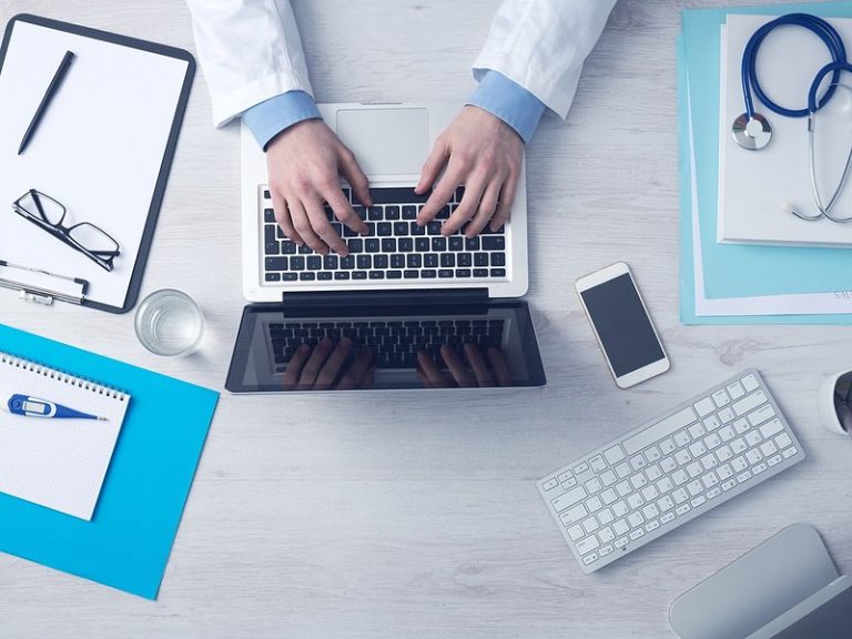 Clinician typing on a laptop at a desk with a stethoscope, clipboard and phone