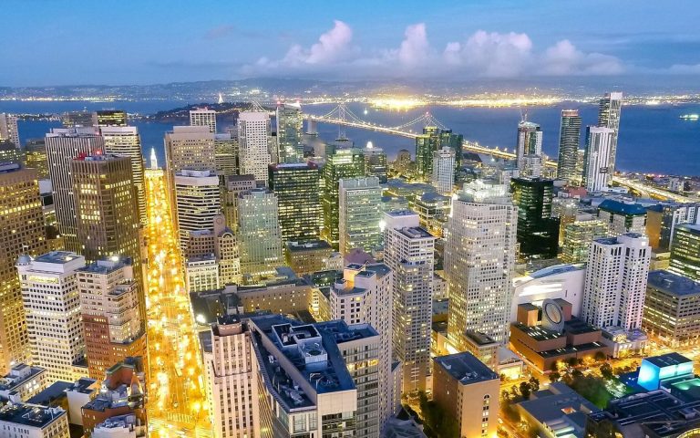 Aerial view of a downtown financial district at dusk with lit streets and a bay bridge