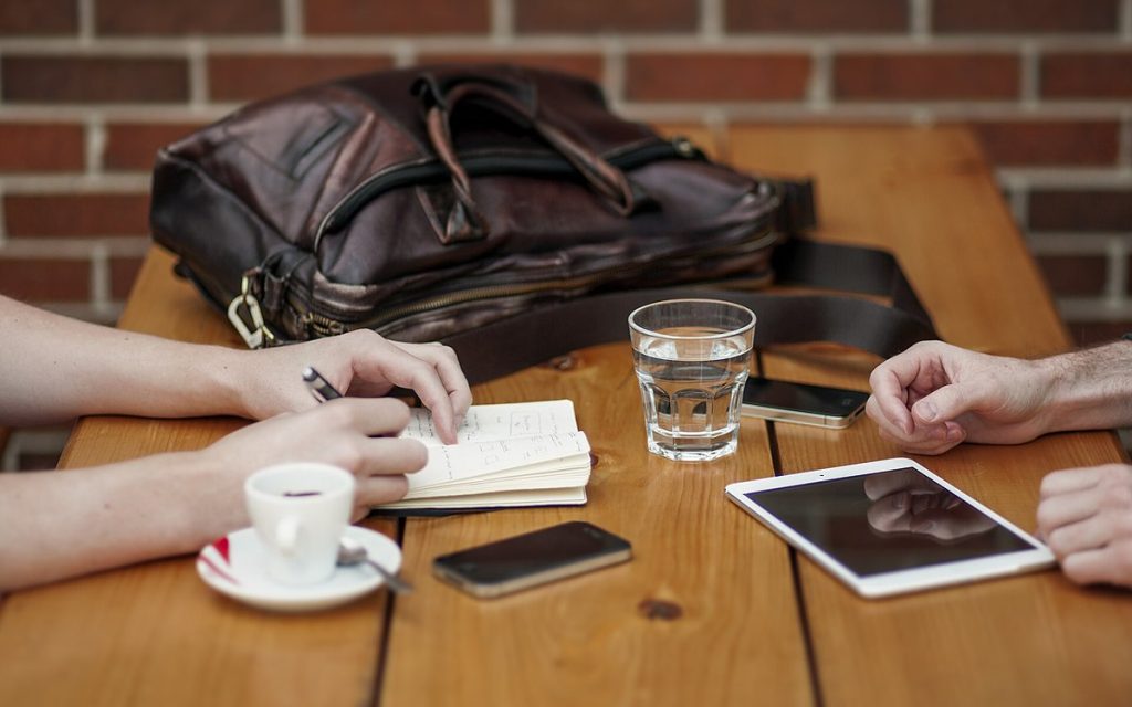 Two colleagues' hands at a wooden table with a tablet, phone, notebook and coffee during a meeting