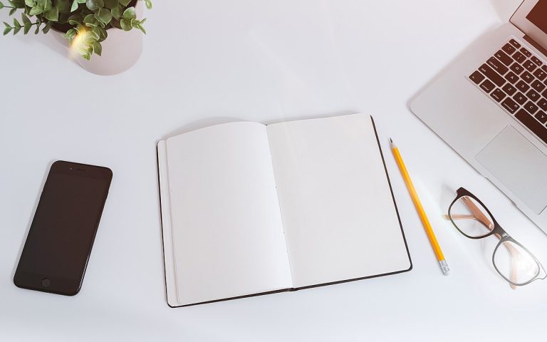 Top-down view of a white desk with an open notebook, pencil, glasses, phone and laptop