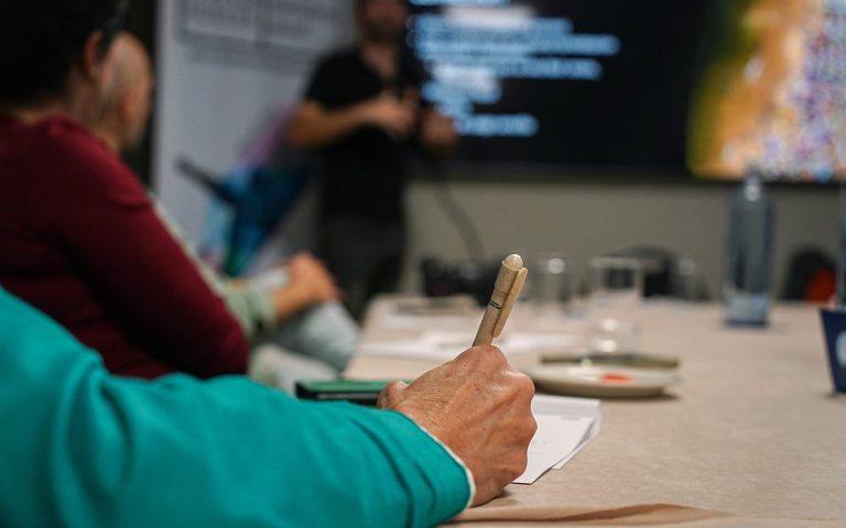 Workshop participant taking notes at a table while a presenter speaks in front of a screen in the background