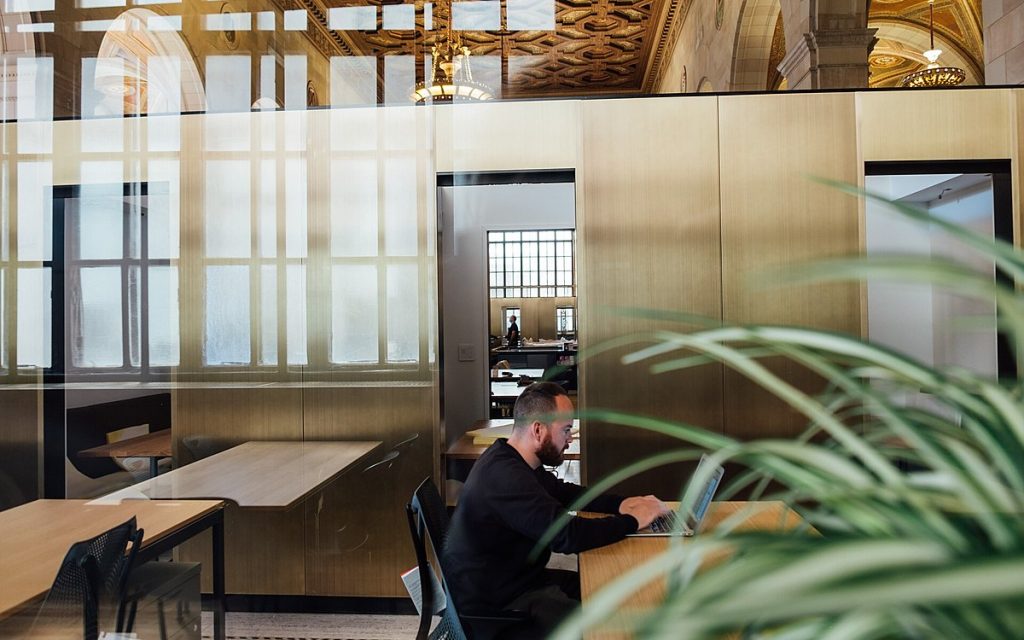 Person working at a laptop in a modern open office with glass partitions and plants