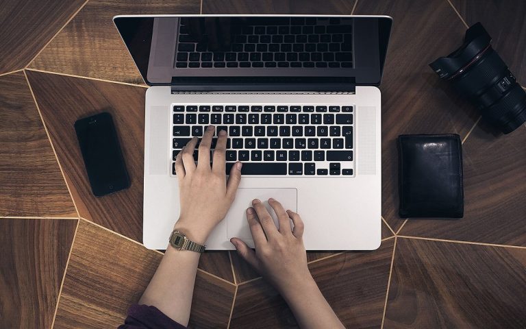Top-down view of hands typing on a laptop on a dark wooden table beside a phone, wallet and camera lens