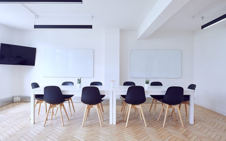 Bright, empty training room with rows of tables, dark chairs, whiteboards and a wall-mounted screen