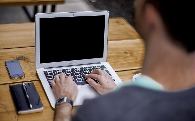 Over-the-shoulder view of a professional typing on a laptop at a wooden desk beside a notebook and phone