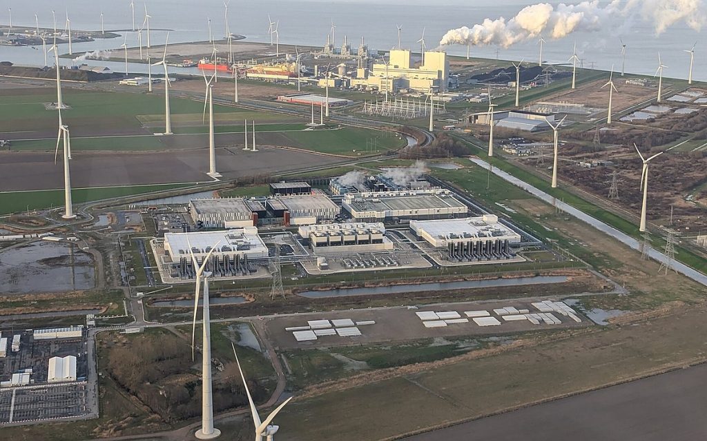 Aerial view of a large data center campus surrounded by wind turbines on flat farmland