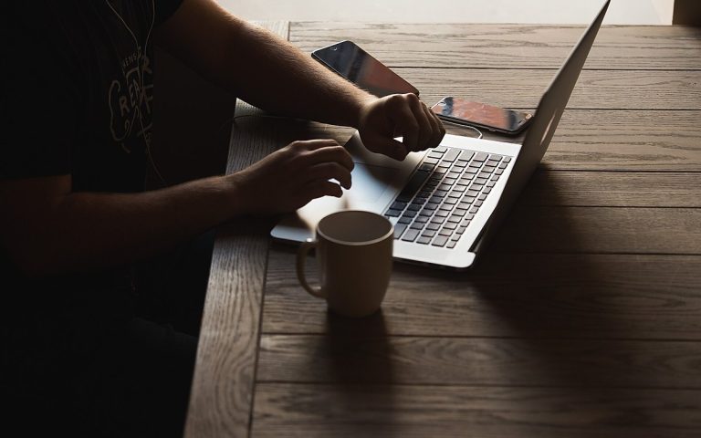 Person in a dim room working on a laptop at a dark wooden table, face out of frame