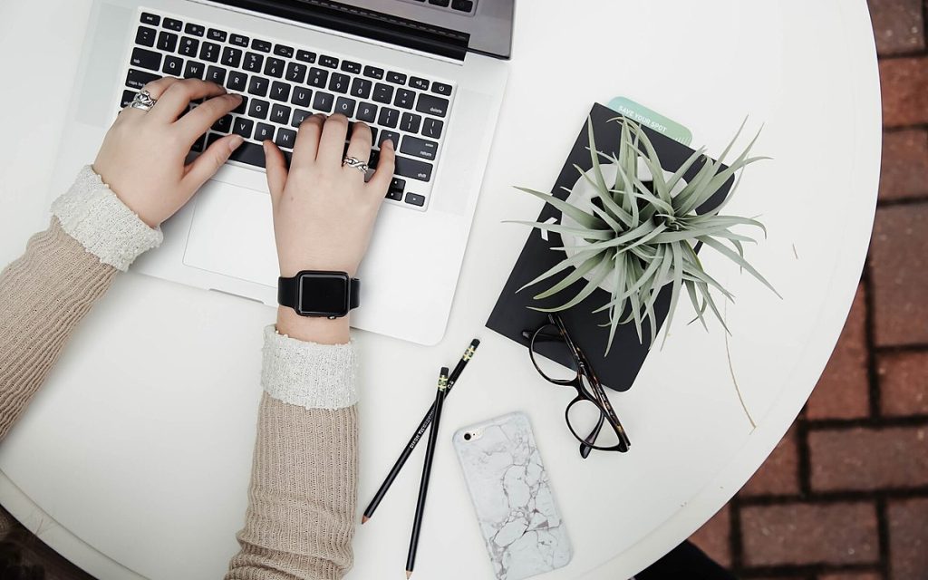 Top-down view of hands typing on a laptop on a white table with a notebook, pencils, glasses and a plant