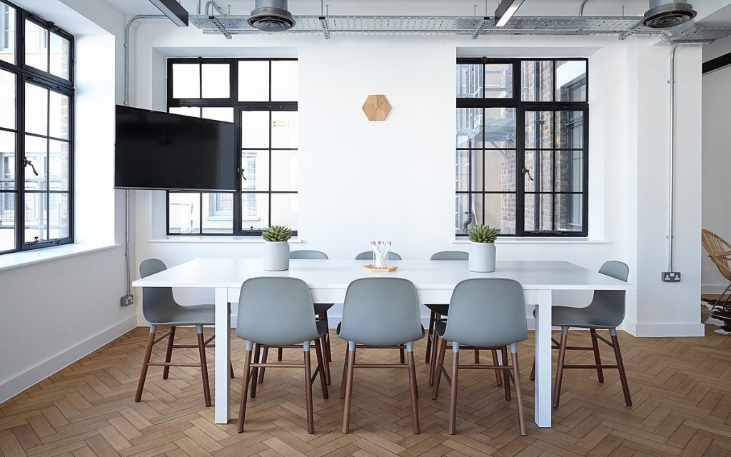 Bright meeting room with a long white table, grey chairs and a wall-mounted screen