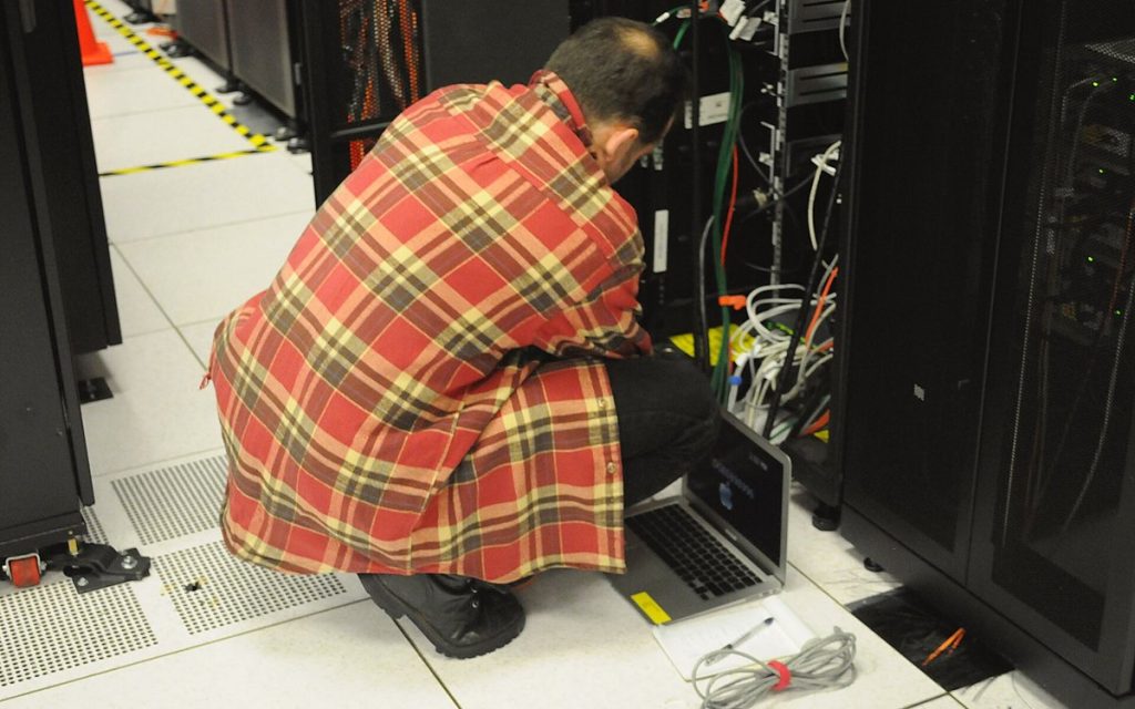 Technician kneeling with a laptop while working on an open server rack in a data center